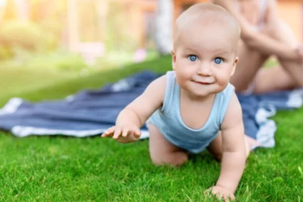 Ein Baby krabbelt auf einem Rasen umher und im Hintergrund sitzt eine Frau auf einer blauen Decke.