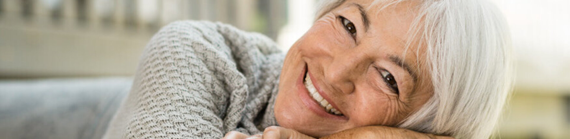 Eine ältere Dame lehnt auf einem Sofa und lacht in die Kamera. 