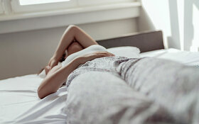 Junge Frau liegt mit starken Regelschmerzen im Bett und hält sich den Bauch.