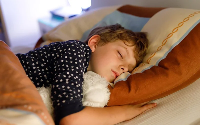 Ein schlafender Junge liegt auf einem Kissen und hält ein weißes Kuscheltier im Arm.