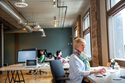 Eine Frau sitzt am Schreibtisch in einem Großraumbüro und arbeitet am Bildschirm.