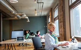 Eine Frau sitzt am Schreibtisch in einem Großraumbüro und arbeitet am Bildschirm.