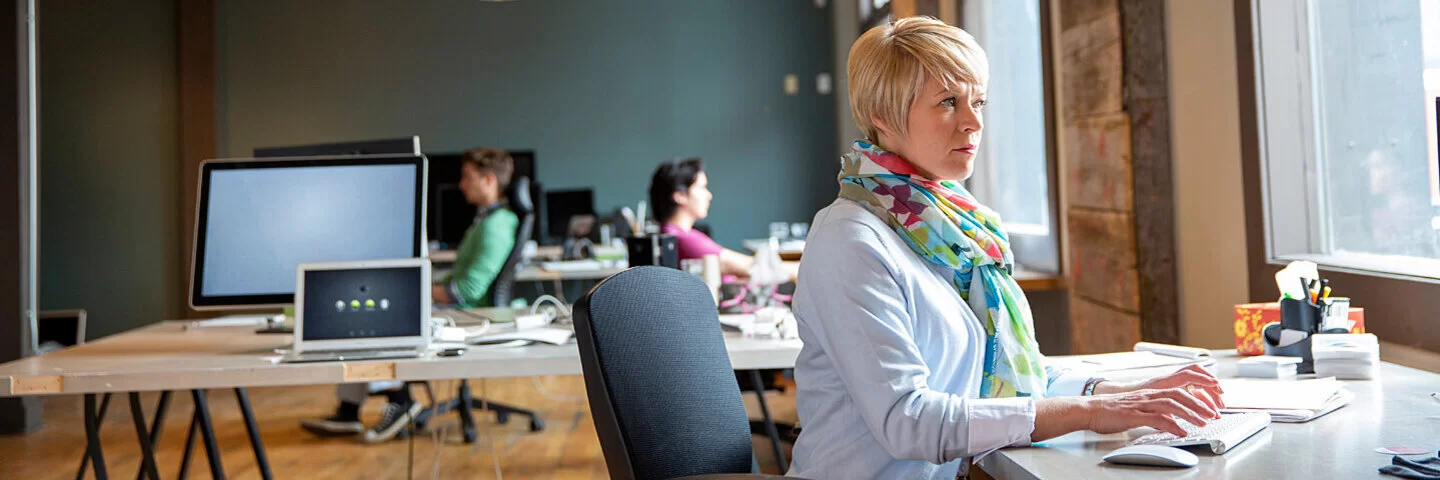 Eine Frau sitzt am Schreibtisch in einem Großraumbüro und arbeitet am Bildschirm.