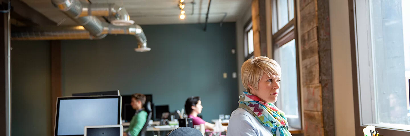 Eine Frau sitzt am Schreibtisch in einem Großraumbüro und arbeitet am Bildschirm.