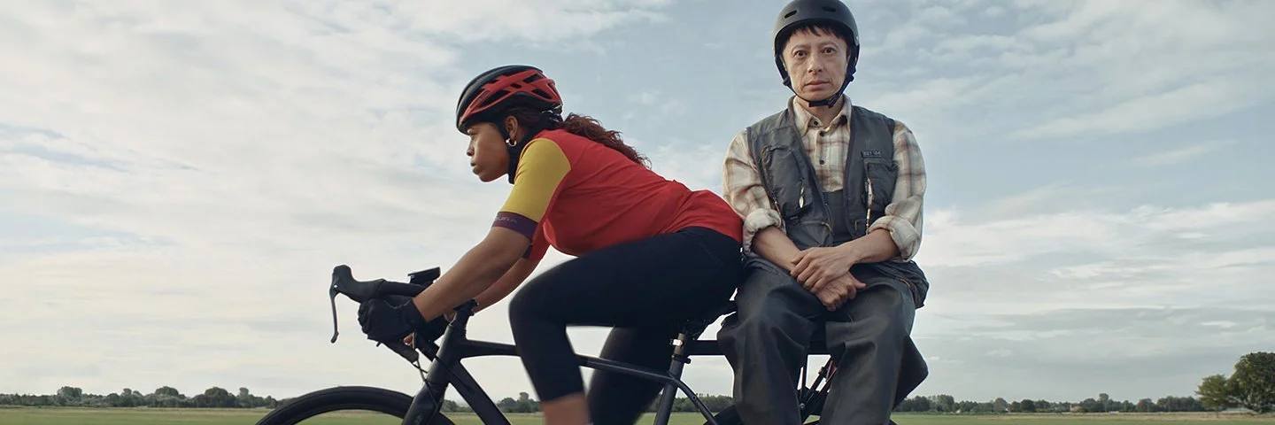Zwei Menschen auf einem Fahrrad, das auf einer Landstraße an einem Feld vorbei fährt.