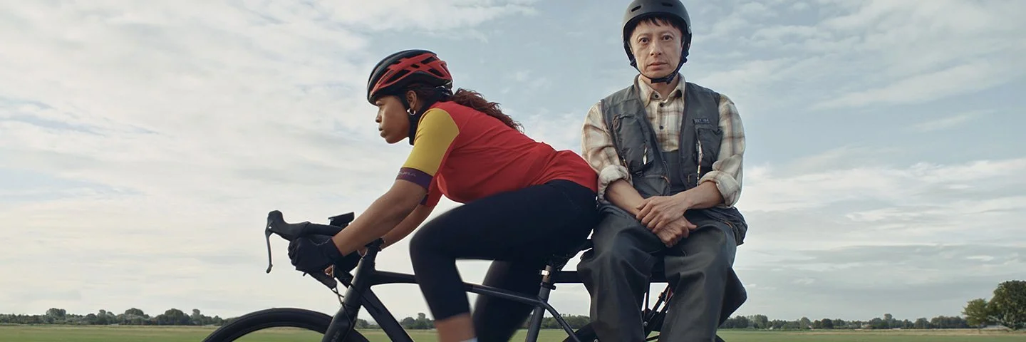 Zwei Menschen auf einem Fahrrad, das auf einer Landstraße an einem Feld vorbei fährt.