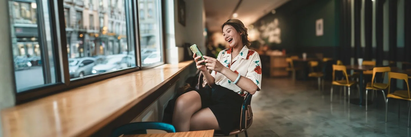 Eine Frau, die in einem Restaurant sitzt und lachend auf ihr Handy schaut.