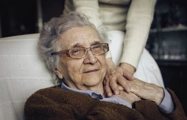 Eine Seniorin liegt auf einem Sessel, eine Person legt fürsorglich ihre Hände auf die Hände der Seniorin