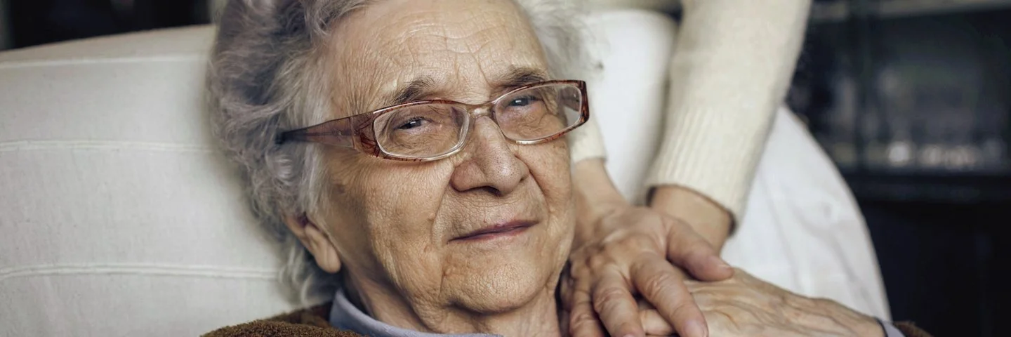 Eine Seniorin liegt auf einem Sessel, eine Person legt fürsorglich ihre Hände auf die Hände der Seniorin