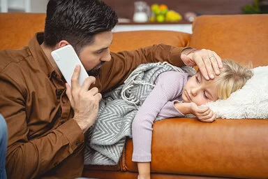 Ein Kind liegt auf einem Sofa. Der Vater hält in der einen Hand einen Telefonhöhrer und fühlt mit der anderen den Kopf des Kindes.