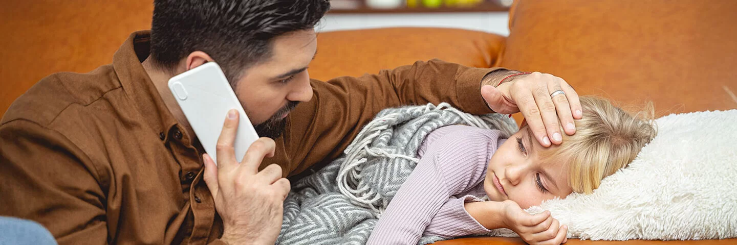 Ein Kind liegt auf einem Sofa. Der Vater hält in der einen Hand einen Telefonhöhrer und fühlt mit der anderen den Kopf des Kindes.