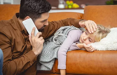 Ein Kind liegt auf einem Sofa. Der Vater hält in der einen Hand einen Telefonhöhrer und fühlt mit der anderen den Kopf des Kindes.