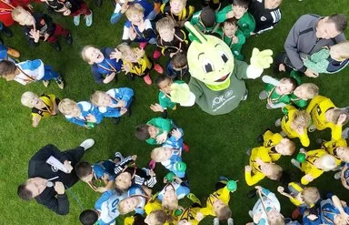 Ein großes Gruppenfoto, von oben aufgenommen, zeigt viele Kinder und einige Erwachsene auf einem Fußballfeld. Im Zentrum steht der grüne Drache Jolinchen (Markenbotschafter für Kindergesundheit der AOK), um den sich die Kinder versammeln. Die Kinder tragen verschiedene Sportkleidungen in Farben wie Blau, Gelb, Rot und Weiß, einige scheinen Medaillen zu tragen.