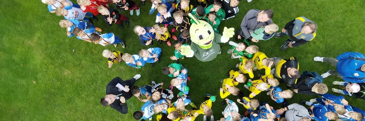 Ein großes Gruppenfoto, von oben aufgenommen, zeigt viele Kinder und einige Erwachsene auf einem Fußballfeld. Im Zentrum steht der grüne Drache Jolinchen (Markenbotschafter für Kindergesundheit der AOK), um den sich die Kinder versammeln. Die Kinder tragen verschiedene Sportkleidungen in Farben wie Blau, Gelb, Rot und Weiß, einige scheinen Medaillen zu tragen.