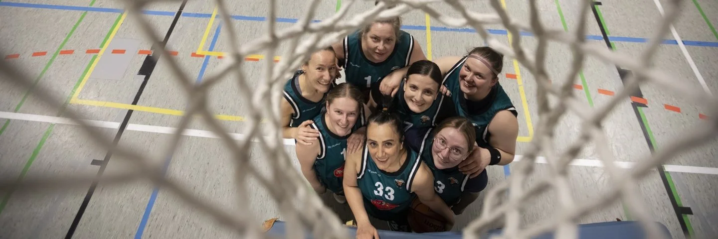 Eine Basketball-Frauenmannschaft steht in einer Sporthalle und blickt von unten durch einen Basketballkorb.