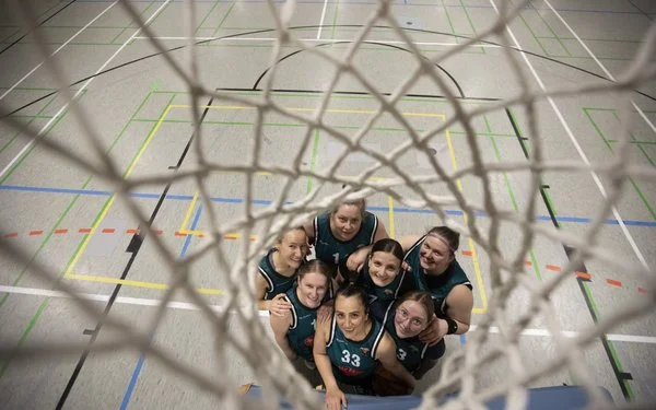 Eine Basketball-Frauenmannschaft steht in einer Sporthalle und blickt von unten durch einen Basketballkorb.
