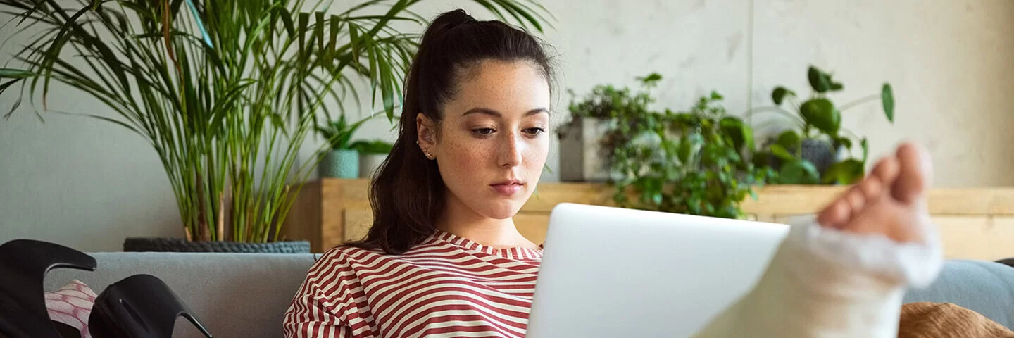 Eine junge Frau sitzt in einem gemütlich eingerichtet Wohnraum auf einer Couch. Ihr rechtes Bein hat einen Gips und ist auf einem Kissen hochgelegt. Auf ihren Beinen balanciert sie einen Laptop, in den sie schaut. Neben ihr auf der Couch liegen zwei Gehstützen.