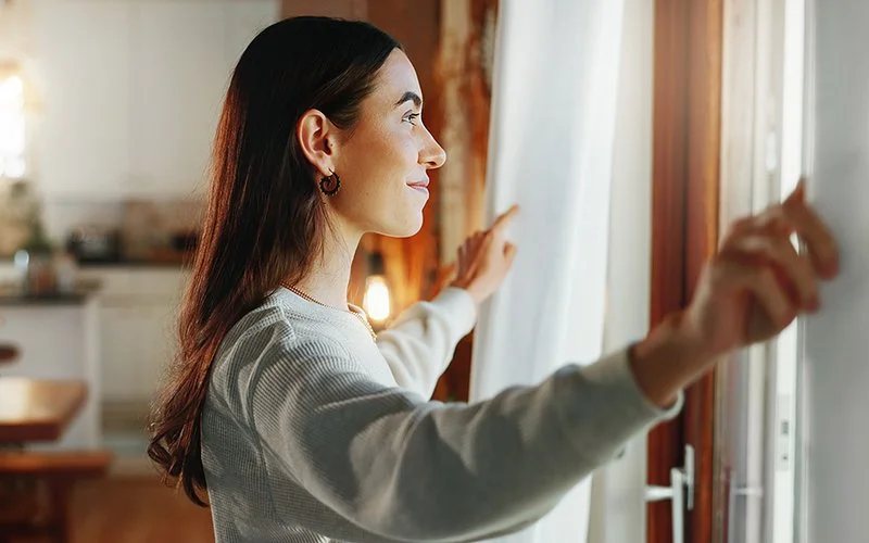 Eine Frau öffnet Gardine und Fenster in einer Küche.