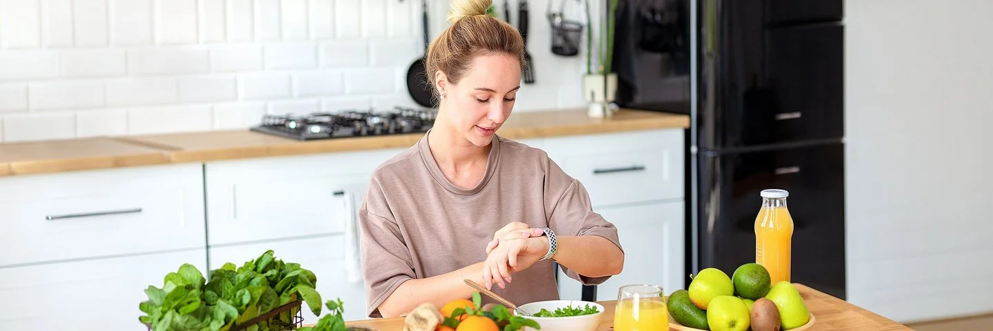 Eine Frau sitzt am Küchentisch und schaut auf ihre Smartwatch. Vor ihr stehen Schalen mit frischem Salat und Obst sowie ein Getränk.
