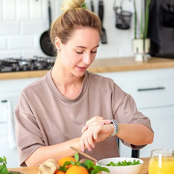 Eine Frau sitzt am Küchentisch und schaut auf ihre Smartwatch. Vor ihr stehen Schalen mit frischem Salat und Obst sowie ein Getränk.