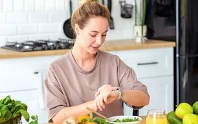 Eine Frau sitzt am Küchentisch und schaut auf ihre Smartwatch. Vor ihr stehen Schalen mit frischem Salat und Obst sowie ein Getränk.