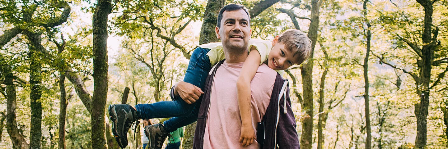 Ein Vater geht im Wald spaziern. Er trägt seinen Sohn über den Rücken geworfen