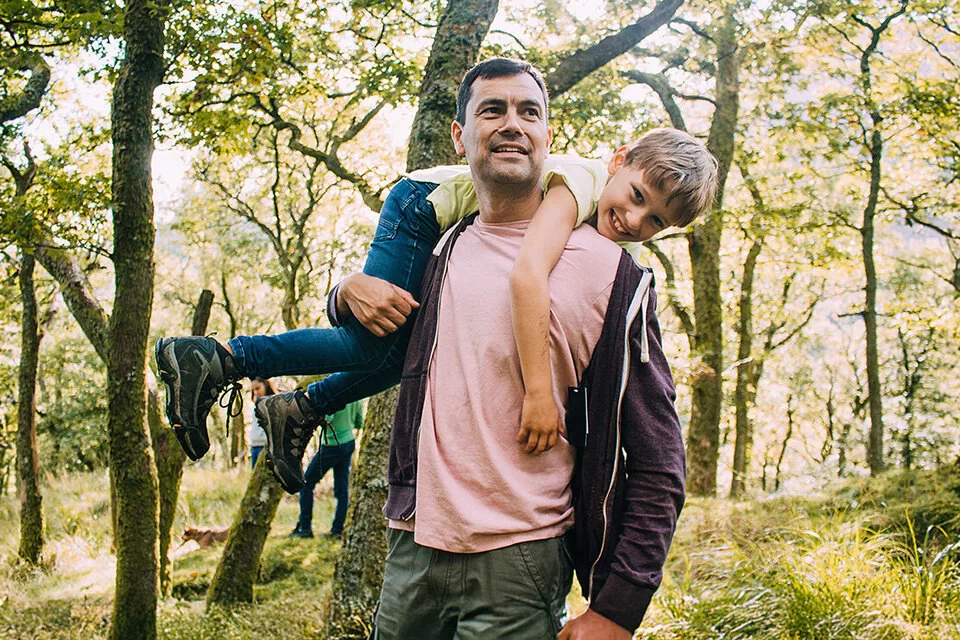 Ein Vater geht im Wald spaziern. Er trägt seinen Sohn über den Rücken geworfen