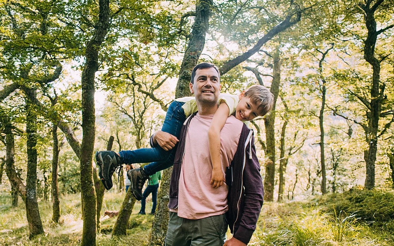 Ein Vater geht im Wald spaziern. Er trägt seinen Sohn über den Rücken geworfen