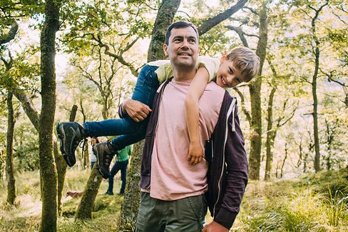 Ein Vater geht im Wald spaziern. Er trägt seinen Sohn über den Rücken geworfen
