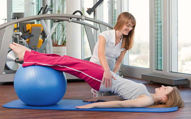 Eine Physiotherapeutin zeigt einer Jugendlichen, die unter Skoliose leidet, Übungen mit dem Gymnastikball.