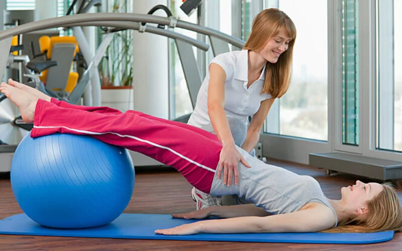 Eine Physiotherapeutin zeigt einer Jugendlichen, die unter Skoliose leidet, Übungen mit dem Gymnastikball.