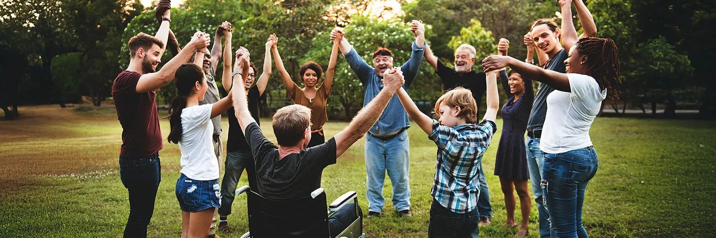 Auf einer Wiese in einem Park stehen im Kreis eine Gruppe von Menschen, darunter Kinder, Jugendliche, junge Erwachsene, Erwachsene und alte Menschen sowie ein Rollstuhlfahrer. Sie fassen sich an den Händen und heben die Arme in die Höhe.