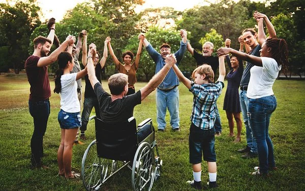 Auf einer Wiese in einem Park stehen im Kreis eine Gruppe von Menschen, darunter Kinder, Jugendliche, junge Erwachsene, Erwachsene und alte Menschen sowie ein Rollstuhlfahrer. Sie fassen sich an den Händen und heben die Arme in die Höhe.