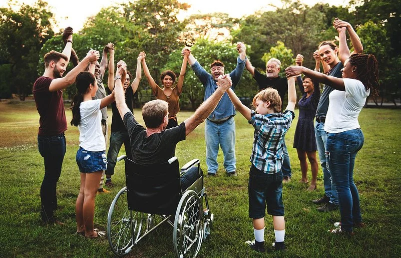 Auf einer Wiese in einem Park stehen im Kreis eine Gruppe von Menschen, darunter Kinder, Jugendliche, junge Erwachsene, Erwachsene und alte Menschen sowie ein Rollstuhlfahrer. Sie fassen sich an den Händen und heben die Arme in die Höhe.