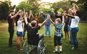 Auf einer Wiese in einem Park stehen im Kreis eine Gruppe von Menschen, darunter Kinder, Jugendliche, junge Erwachsene, Erwachsene und alte Menschen sowie ein Rollstuhlfahrer. Sie fassen sich an den Händen und heben die Arme in die Höhe.