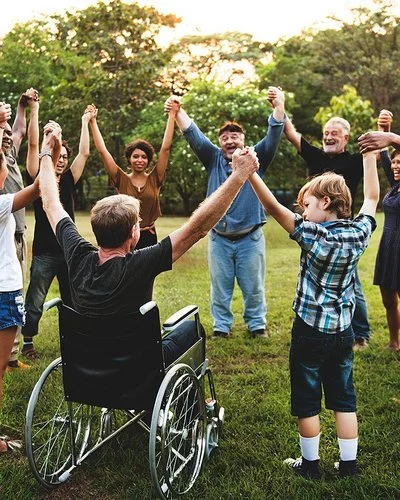 Auf einer Wiese in einem Park stehen im Kreis eine Gruppe von Menschen, darunter Kinder, Jugendliche, junge Erwachsene, Erwachsene und alte Menschen sowie ein Rollstuhlfahrer. Sie fassen sich an den Händen und heben die Arme in die Höhe.