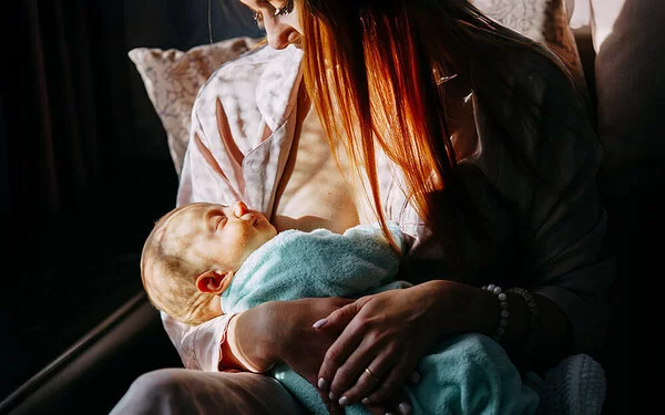 Stillende Mutter in einem hellen Morgenlicht, die ihr in eine Decke gewickeltes, schlafendes Baby an der Brust hält.