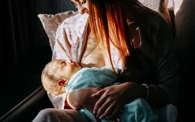 Stillende Mutter in einem hellen Morgenlicht, die ihr in eine Decke gewickeltes, schlafendes Baby an der Brust hält.