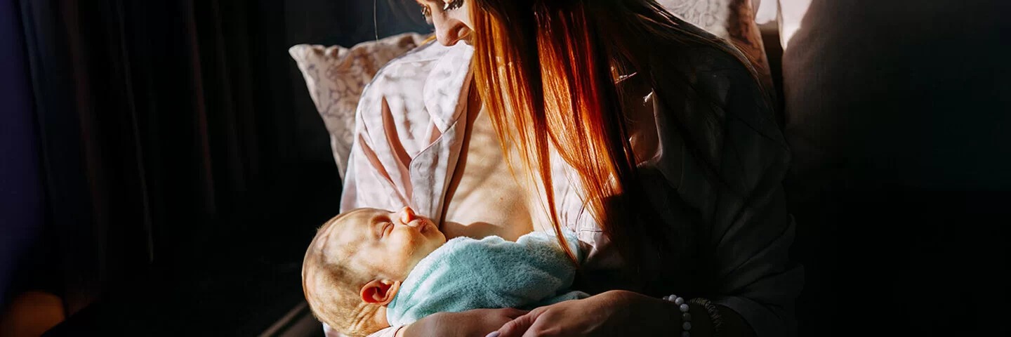 Stillende Mutter in einem hellen Morgenlicht, die ihr in eine Decke gewickeltes, schlafendes Baby an der Brust hält.