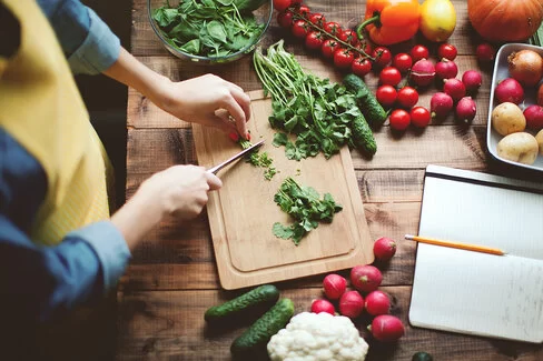 Person schneidet frischen Koriander auf einem Holzbrett, auf dem Tisch liegen Tomaten, Radieschen und weiteres Gemüse. 