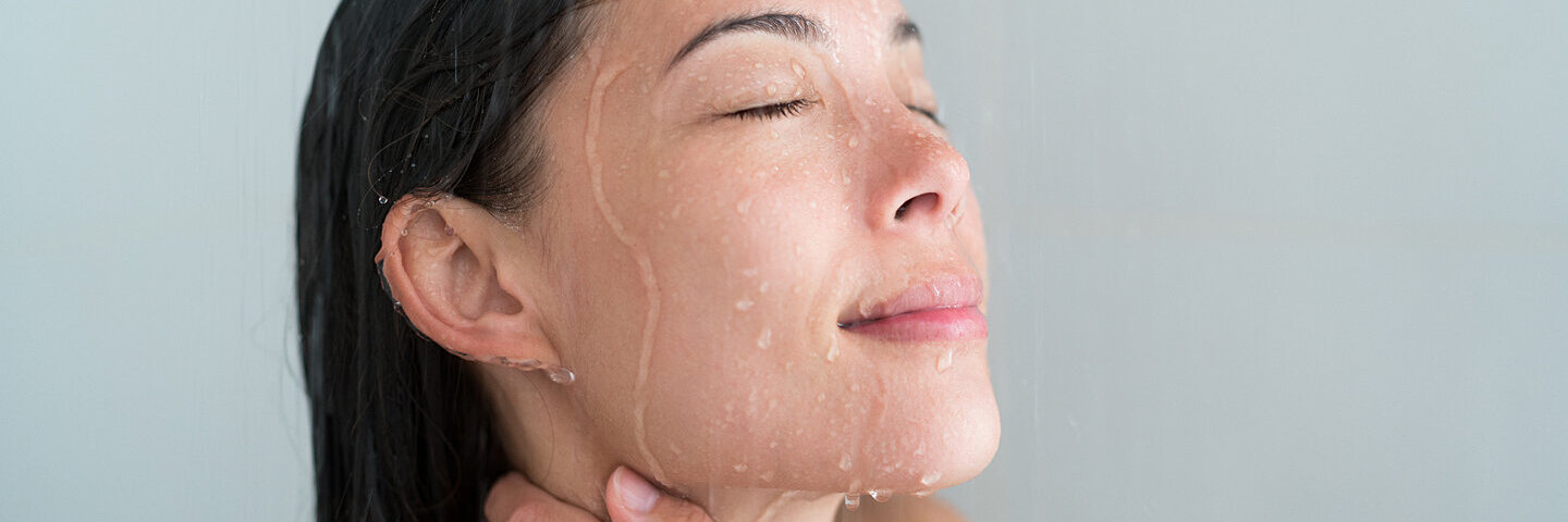 Eine junge Frau lässt unter der Dusche Wasser über ihr Gesicht laufen.