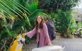 Ein junge Touristin mit Rucksack, die hinter einem gelben Motorroller steht.