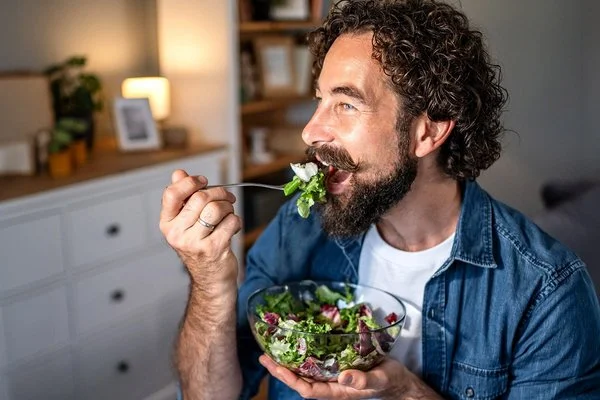 Ein Mann mit Vollbart, offenem Jeanshemd und weißem T-Shirt isst gut gelaunt einen Salat aus einer Glasschüssel.