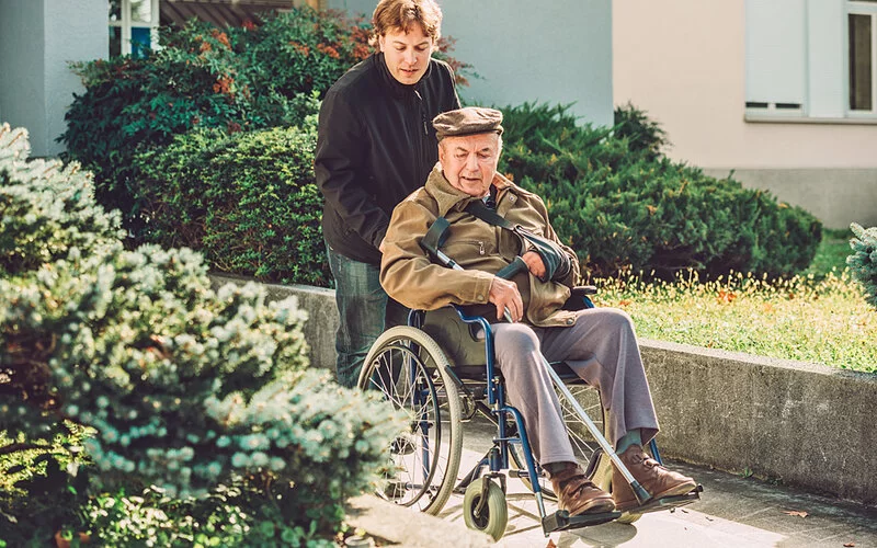 Ein alter Mann wird von einem jungen Angehörigen im Rollstuhl einen Gehweg entlanggeschoben.