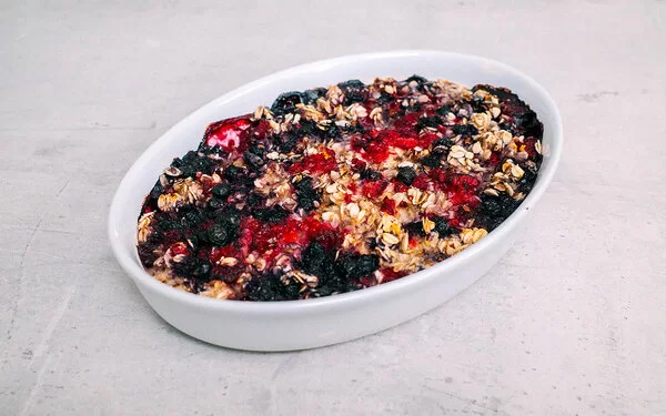 Baked Oatmeal mit Beeren nach einem Rezept von Felicitas Then, in einer Auflaufform auf einer grauen Arbeitsplatte. Im Hintergrund herzhafte Hafer-Cookies mit Bergkäse.