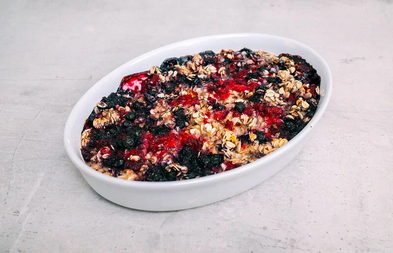 Baked Oatmeal mit Beeren nach einem Rezept von Felicitas Then, in einer Auflaufform auf einer grauen Arbeitsplatte. Im Hintergrund herzhafte Hafer-Cookies mit Bergkäse.