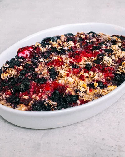 Baked Oatmeal mit Beeren nach einem Rezept von Felicitas Then, in einer Auflaufform auf einer grauen Arbeitsplatte. Im Hintergrund herzhafte Hafer-Cookies mit Bergkäse.