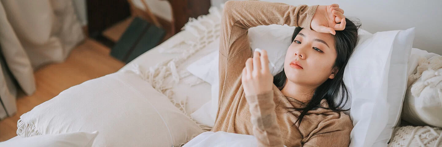 Eine Frau liegt im Bett und hält ein Fieberthermometer in der Hand.