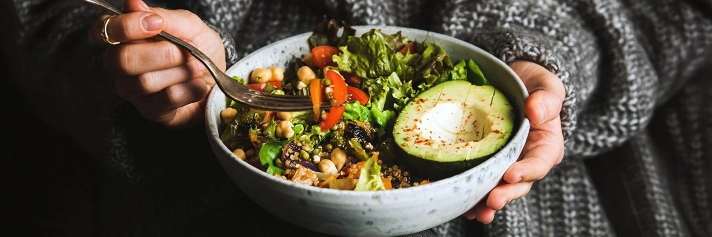 Eine nicht erkennbare Person in einem weiten grauen Strickpullover hält mit der linken Hand eine Schale mit buntem Salat, Avocadostücken, Tomaten und Kichererbsen auf dem Schoß. In der rechten Hand hält sie eine Gabel mit Tomatenstücken.