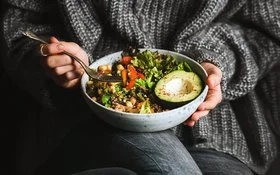Eine nicht erkennbare Person in einem weiten grauen Strickpullover hält mit der linken Hand eine Schale mit buntem Salat, Avocadostücken, Tomaten und Kichererbsen auf dem Schoß. In der rechten Hand hält sie eine Gabel mit Tomatenstücken.